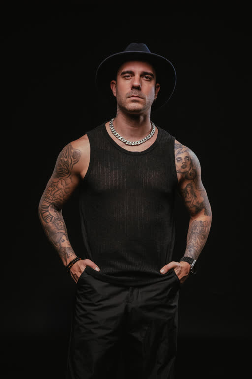 Juanjo Vivancos posando frontalmente, cadena de plata gruesa y sombrero negro, sobre fondo negro estudio.